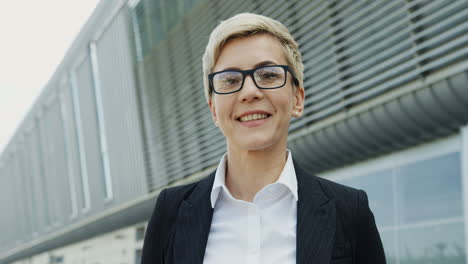 Portrait-of-the-succesful-middle-aged-businesswoman-with-short-blond-hair-turning-her-head-to-the-camera-and-smiling-on-the-big-urban-building-background.-Close-up